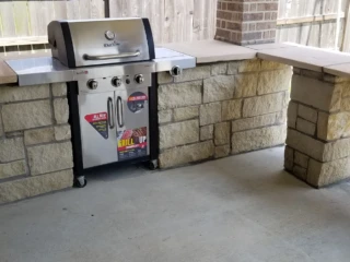 A stainless steel gas grill is built into a stone outdoor kitchen on a covered patio with a concrete floor.