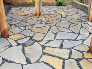 Stone patio with irregular-shaped pavers, bordered by gravel, wooden posts, and tall planters with green plants.