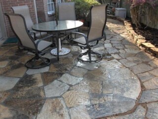 Outdoor patio with irregular stone flooring, a round glass table, and four chairs near a brick wall and garden bed.